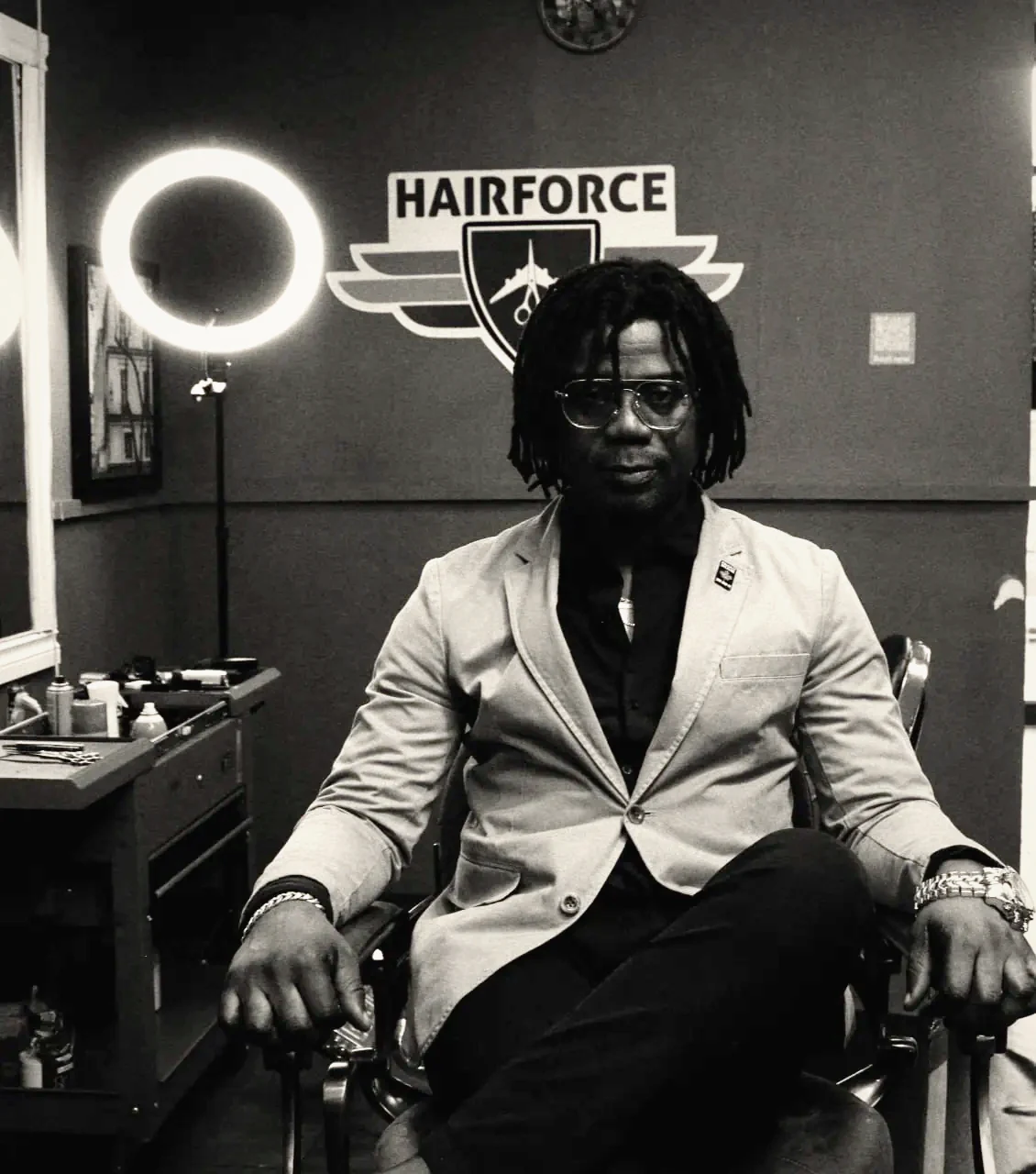 The HAIRFORCE master barber seated in a barber chair beneath the illuminated HAIRFORCE sign